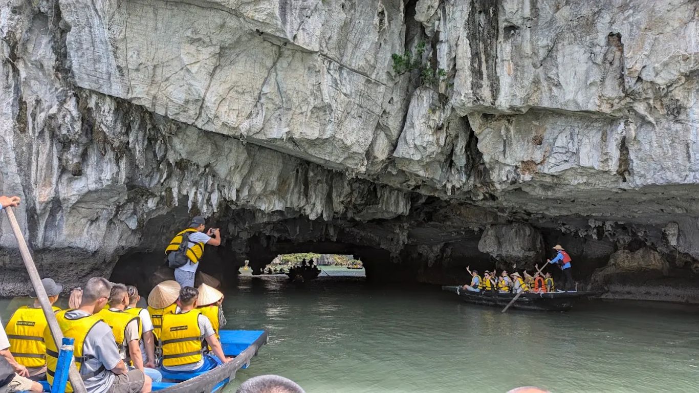 Tourists on wooden bamboo rowing boats entering Ba Hang Cave through a narrow limestone archway in Halong Bay, guided by local rowers in conical hats.