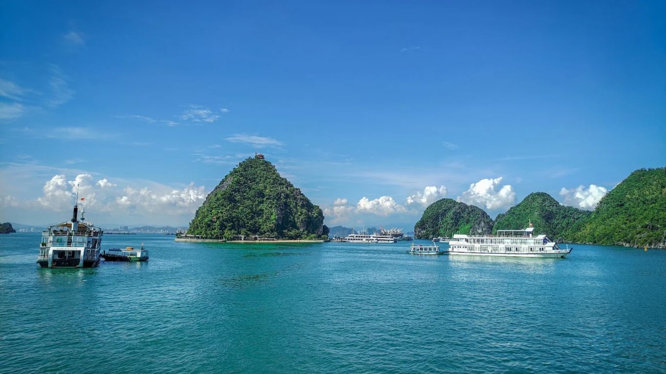Ti Top Island in Halong Bay on a clear sunny day, with a green limestone peak rising from turquoise water, cruise ships anchored nearby and karst islands stretching into the distance.