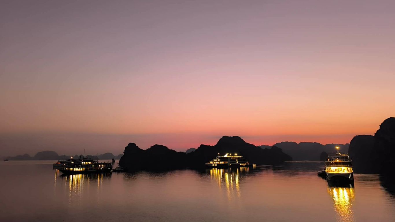 Cruise ships anchored in Halong Bay at sunset, their warm lights reflecting on calm water beneath a pink and purple evening sky silhouetted by limestone karsts.